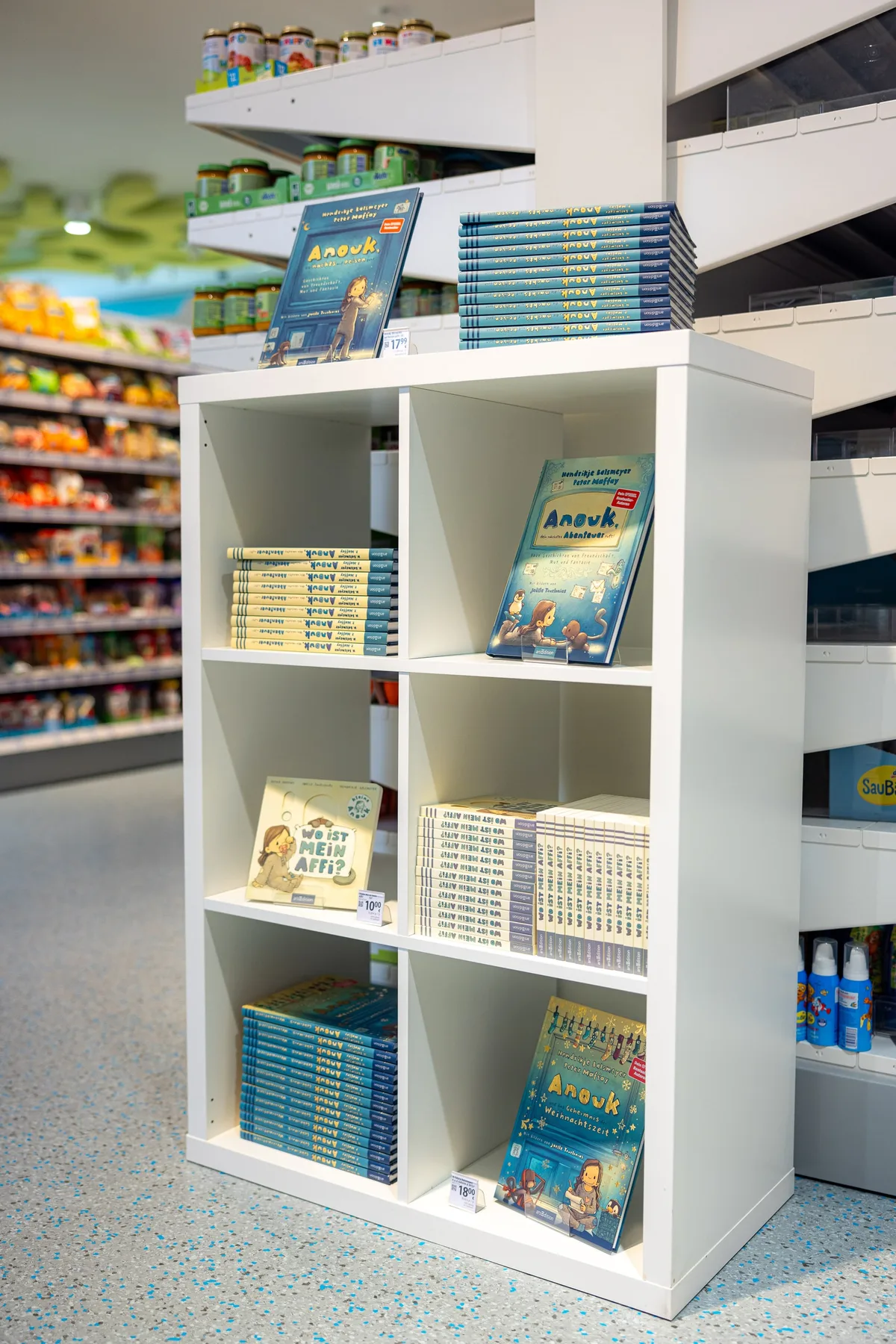 Weißes Regal mit gestapelten Kinderbüchern und aufgestellten Covern, dahinter Lebensmittelregale im Drogeriemarkt