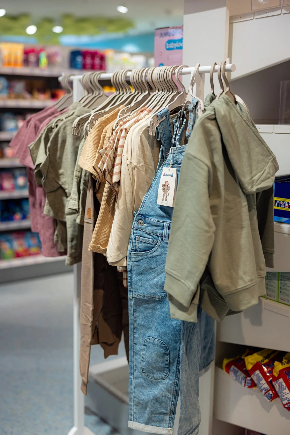 Kleiderstange mit Kinderkleidung in Grün, Beige und Rosa, vorne eine Jeans-Latzhose mit Papieranhänger