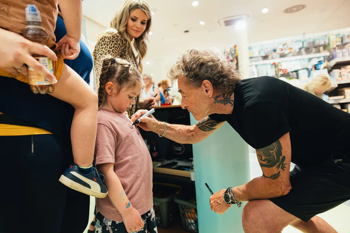 Peter Maffay beugt sich mit einem Filzstift zu einem Mädchen im rosa Shirt, dahinter eine Frau im Leopardenmantel