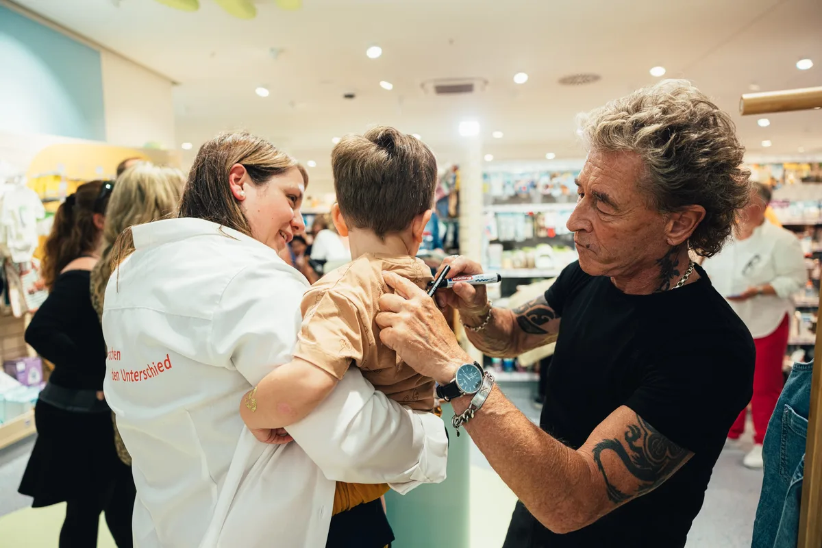 Peter Maffay signiert mit Filzstift den Rücken eines Jungen, den eine Frau in weißer Jacke auf dem Arm trägt