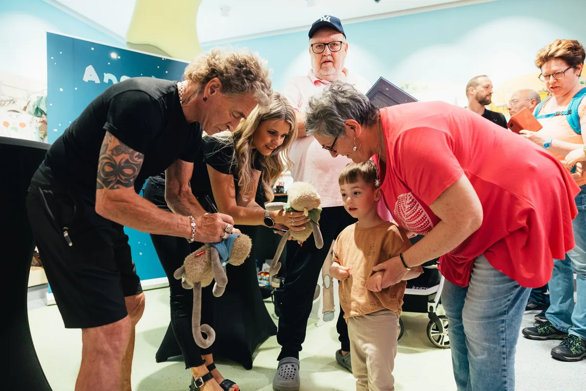 Peter Maffay signiert ein Plüschäffchen, daneben eine lächelnde Frau, davor ein Kleinkind und eine Frau im roten Shirt