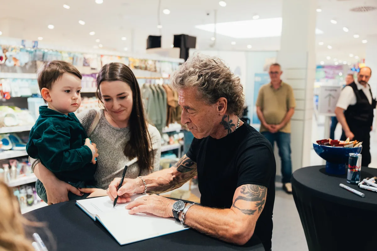 Peter Maffay signiert ein Buch, daneben ein Kleinkind in grünem Samtjackett auf dem Arm einer Frau