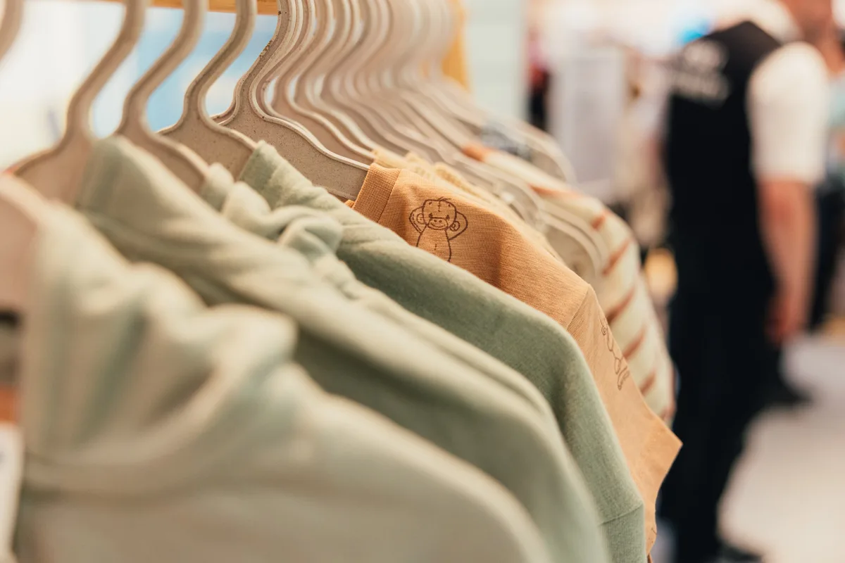 Unscharfe Kleiderstange mit Hoodies und orangem Shirt mit Äffchen-Motiv, dahinter eine Person in Weste
