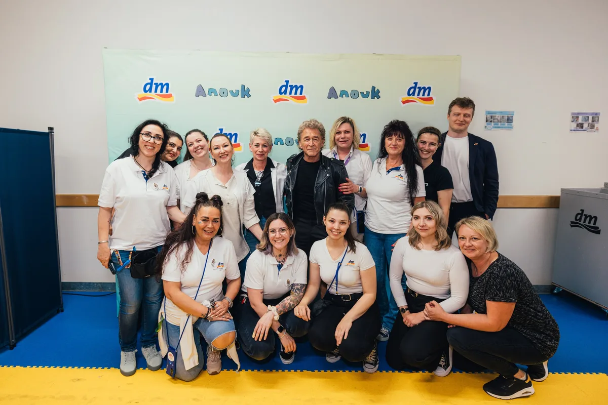 Gruppenbild vor einer Anouk-Wand: Mann in Lederjacke in der Mitte, umringt von Beschäftigten in weißen Shirts