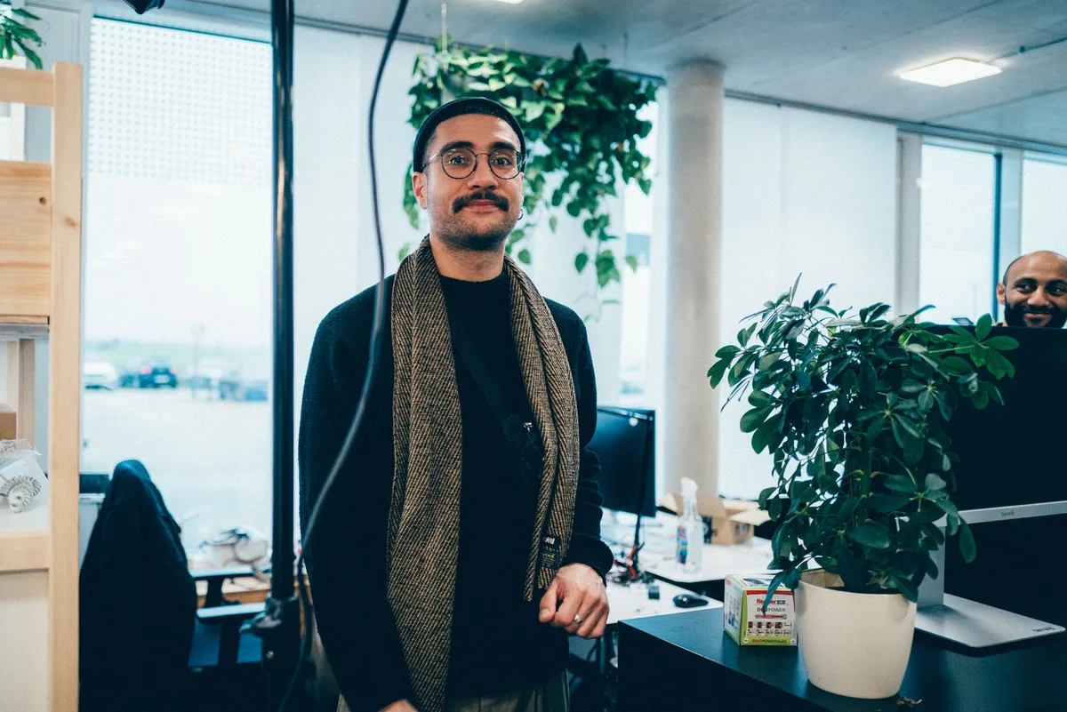 Mann mit Mütze, runder Brille, Schnurrbart und gestreiftem Schal steht neben einer Zimmerpflanze im Büro
