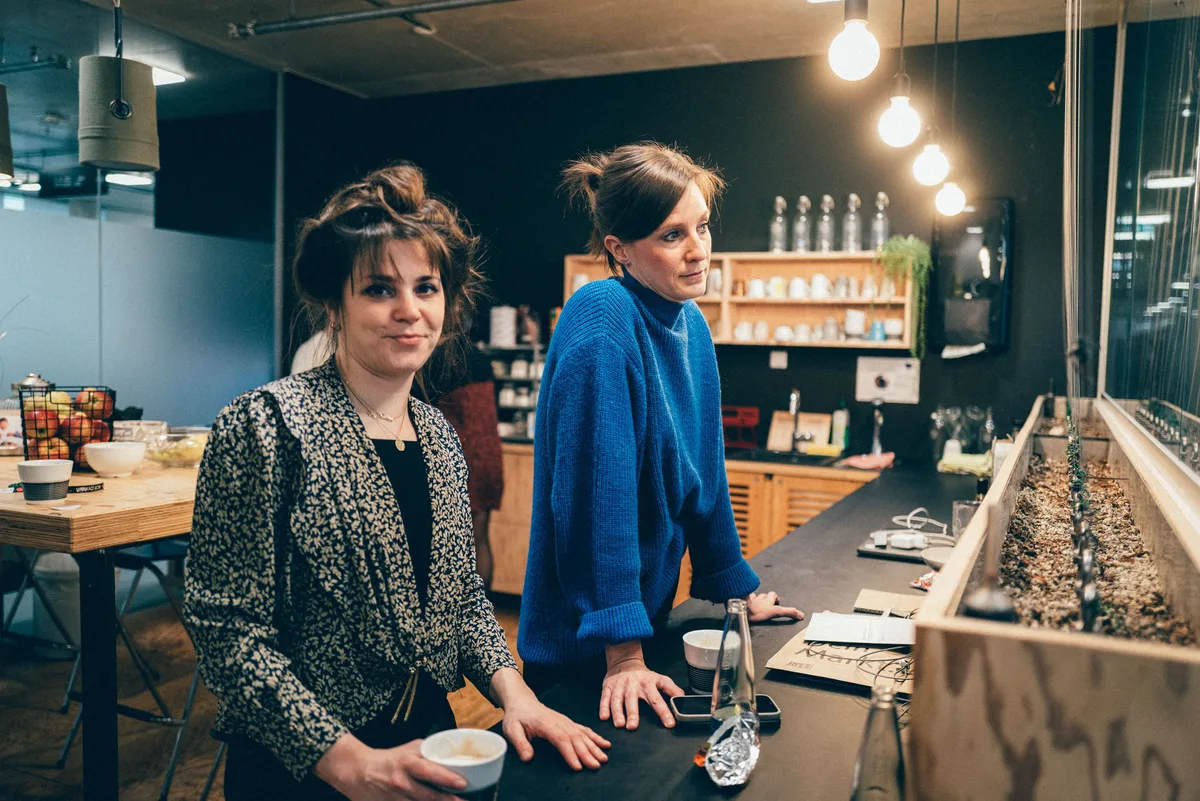 Zwei Frauen an der Küchentheke, links im gemusterten Blazer mit Tasse, rechts im blauen Pullover, dahinter Regale