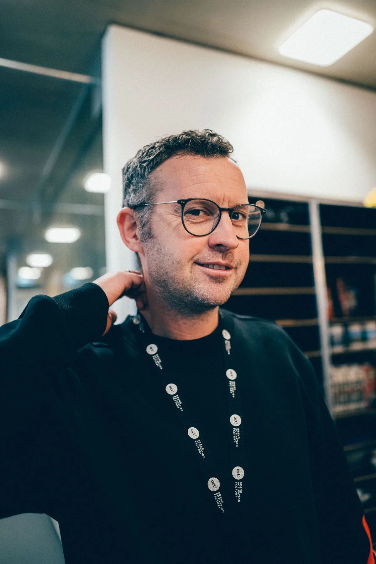 Mann mit Brille und Dreitagebart, Lanyard um den Hals, fasst sich im Hochformat mit einer Hand in den Nacken