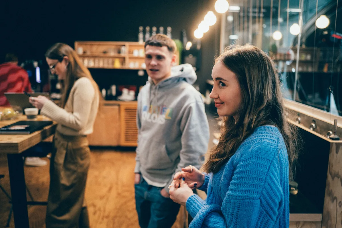 Frau im blauen Strickpulli blickt seitlich aus dem Bild, hinter ihr ein junger Mann im grauen Hoodie und eine Frau am Tisch