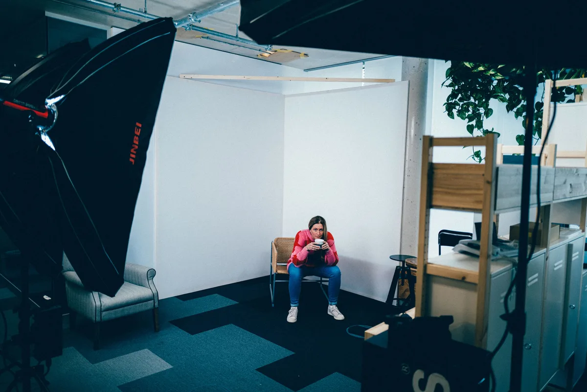Frau im rosa Pulli sitzt mit einer Tasse auf einem Korbstuhl vor den Stellwänden, links ragt eine Softbox ins Bild