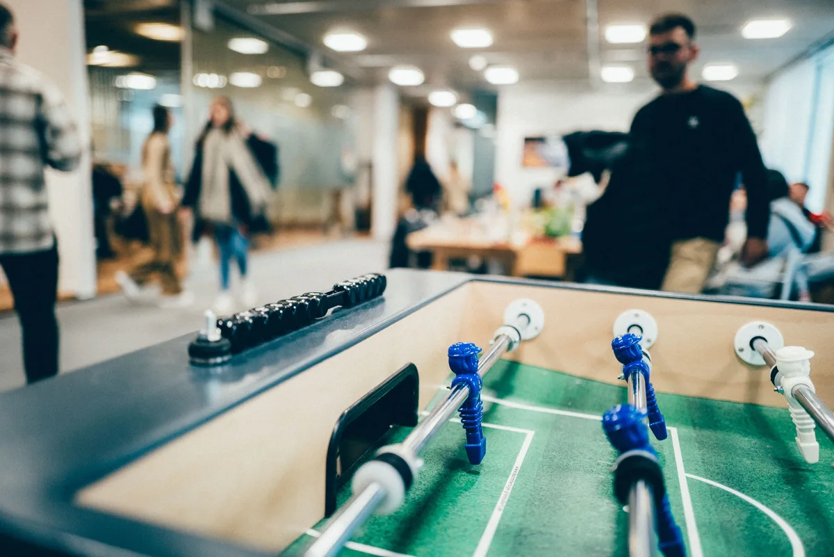 Kicker mit blauen Figuren im Vordergrund, unscharf dahinter Menschen im Büro