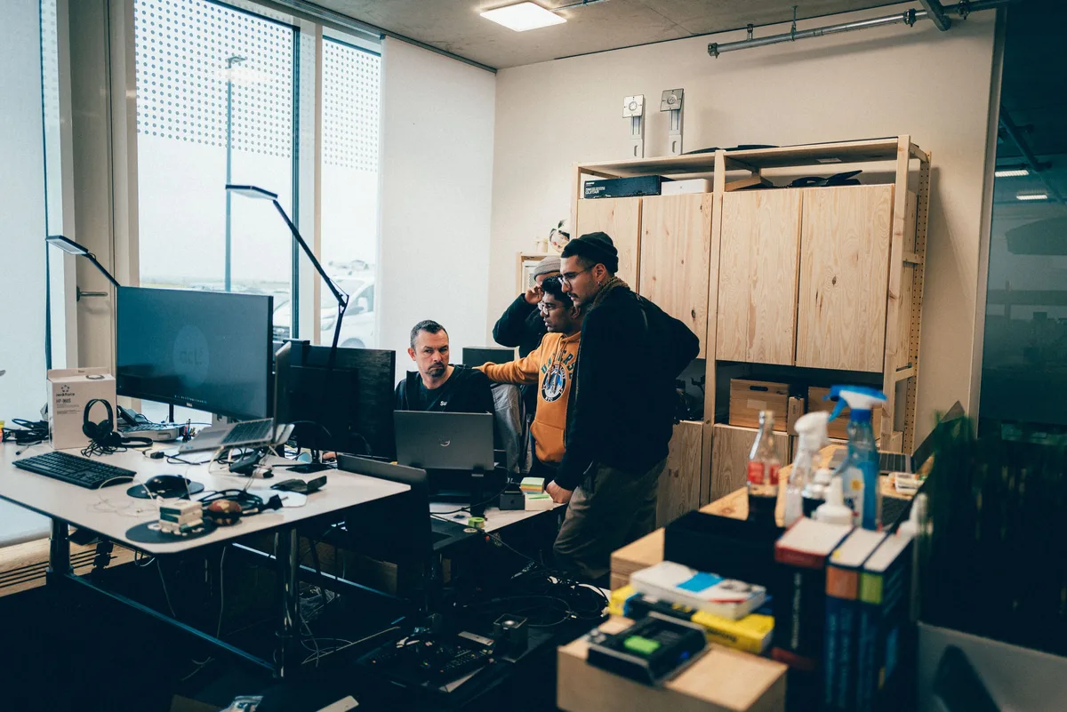 Vier Männer an einem Schreibtisch vor Monitoren, einer sitzt am Laptop, die anderen stehen dahinter und schauen zu