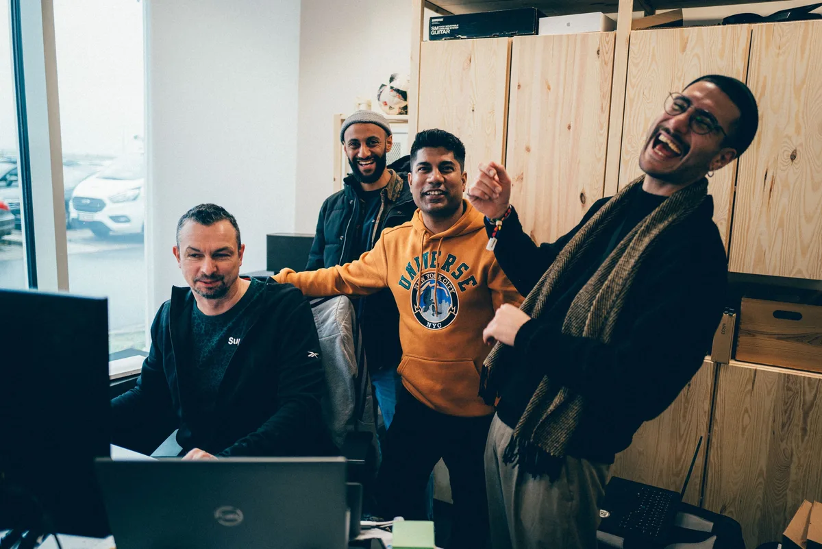 Vier Männer lachen am Schreibtisch, rechts einer mit Schal und Brille mit weit offenem Mund, links einer am Laptop