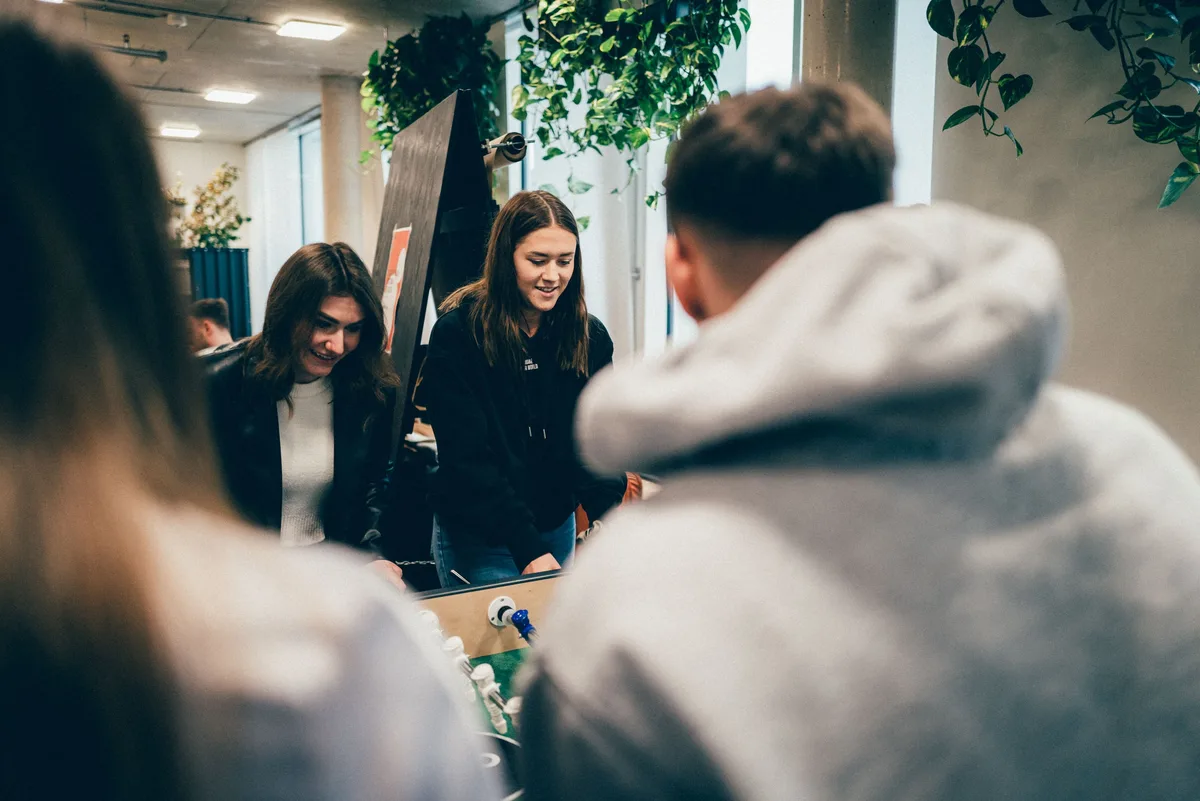 Zwei Frauen lachen am Kicker, rechts verdeckt der Rücken eines Mannes im Hoodie halb das Bild, dahinter Zimmerpflanzen