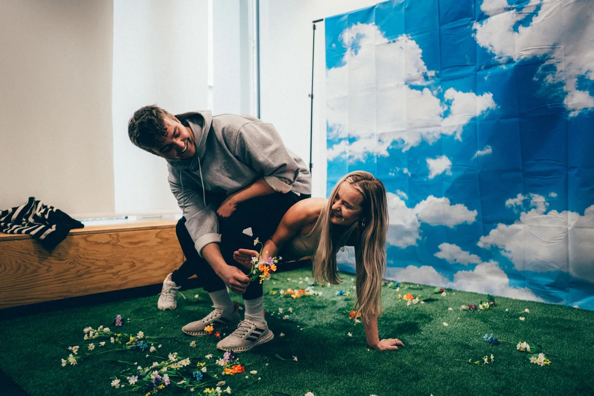 Dicht dran: Mann beugt sich lachend über die Frau auf Kunstrasen, beide halten Blumen, rechts die Wolkenkulisse
