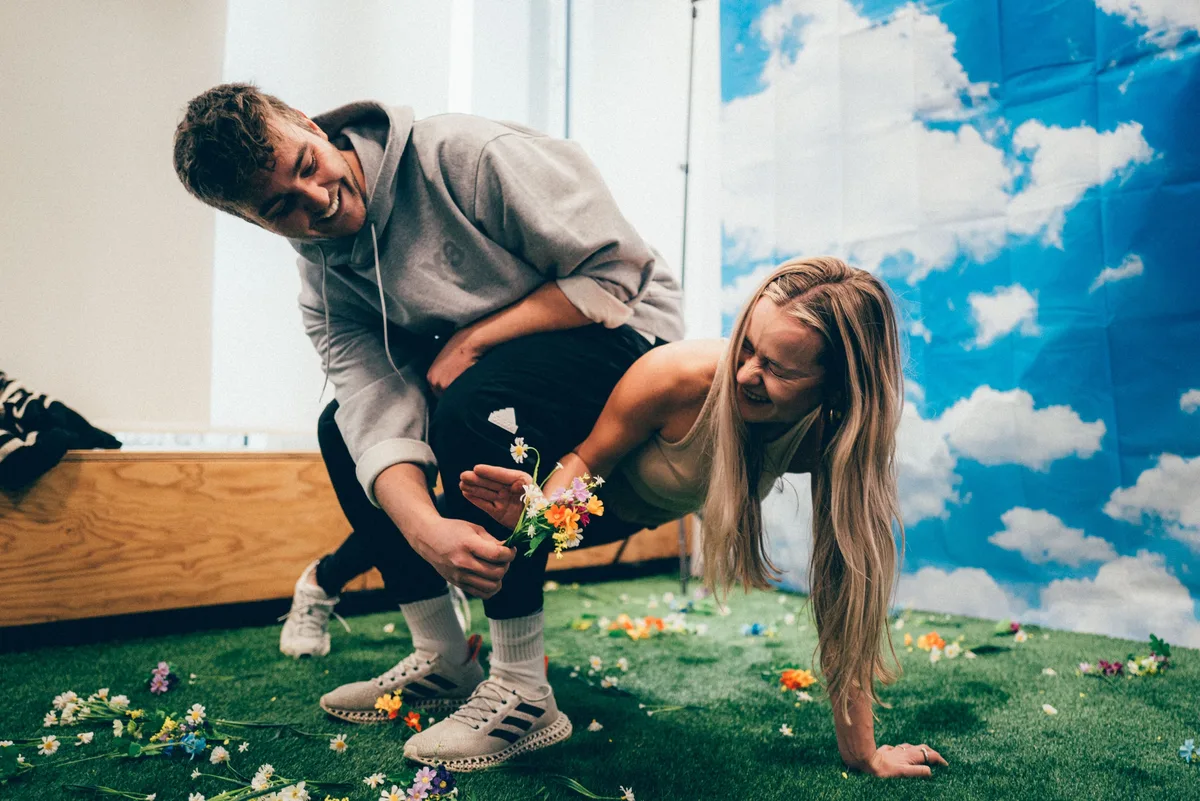 Beide lachen herzhaft, der Mann hält einen Blumenstrauß, auf dem Kunstrasen liegen verstreute Blumen