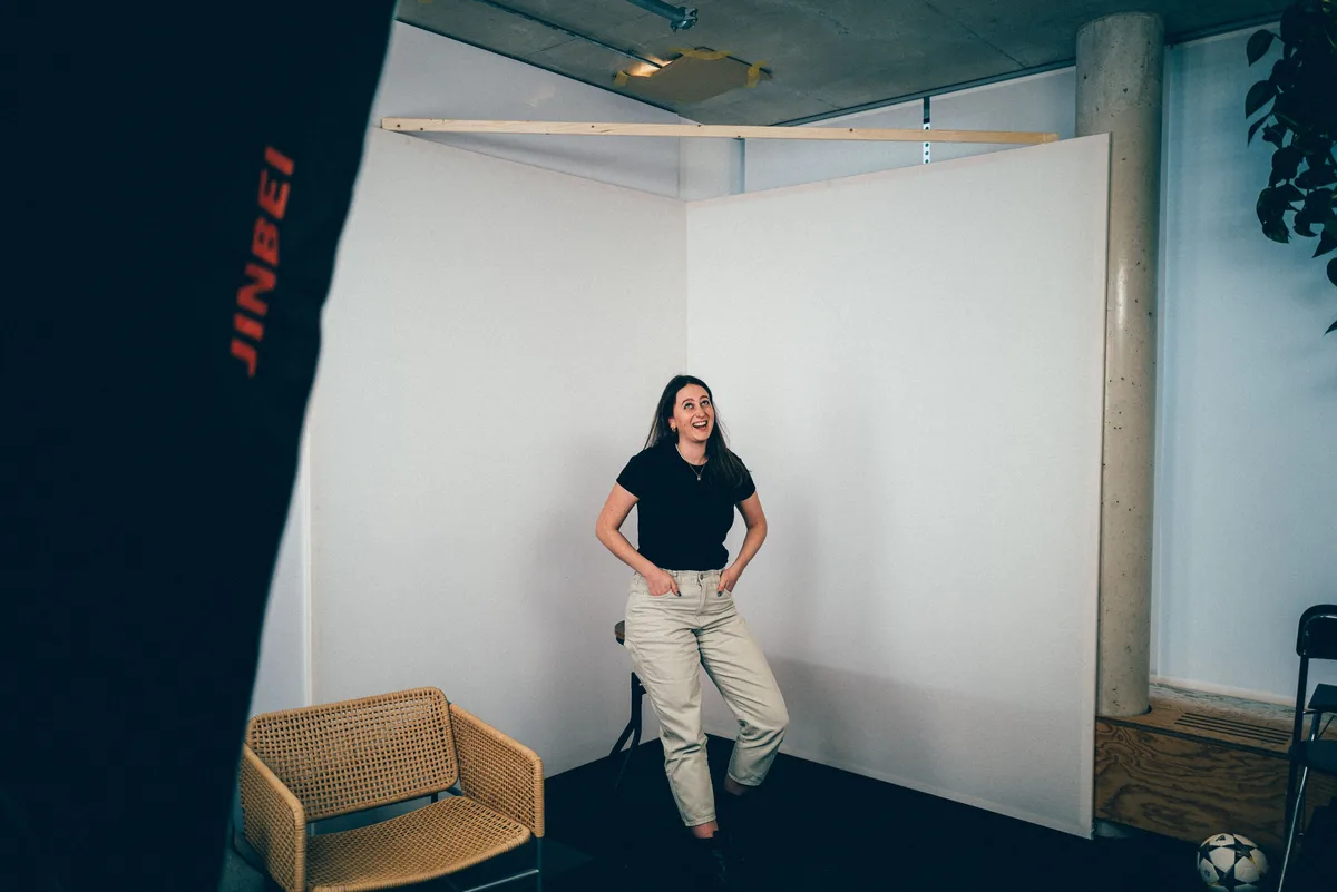 Frau im schwarzen Shirt lehnt lässig vor weißen Stellwänden und lacht nach oben, links eine Softbox, rechts ein Fußball