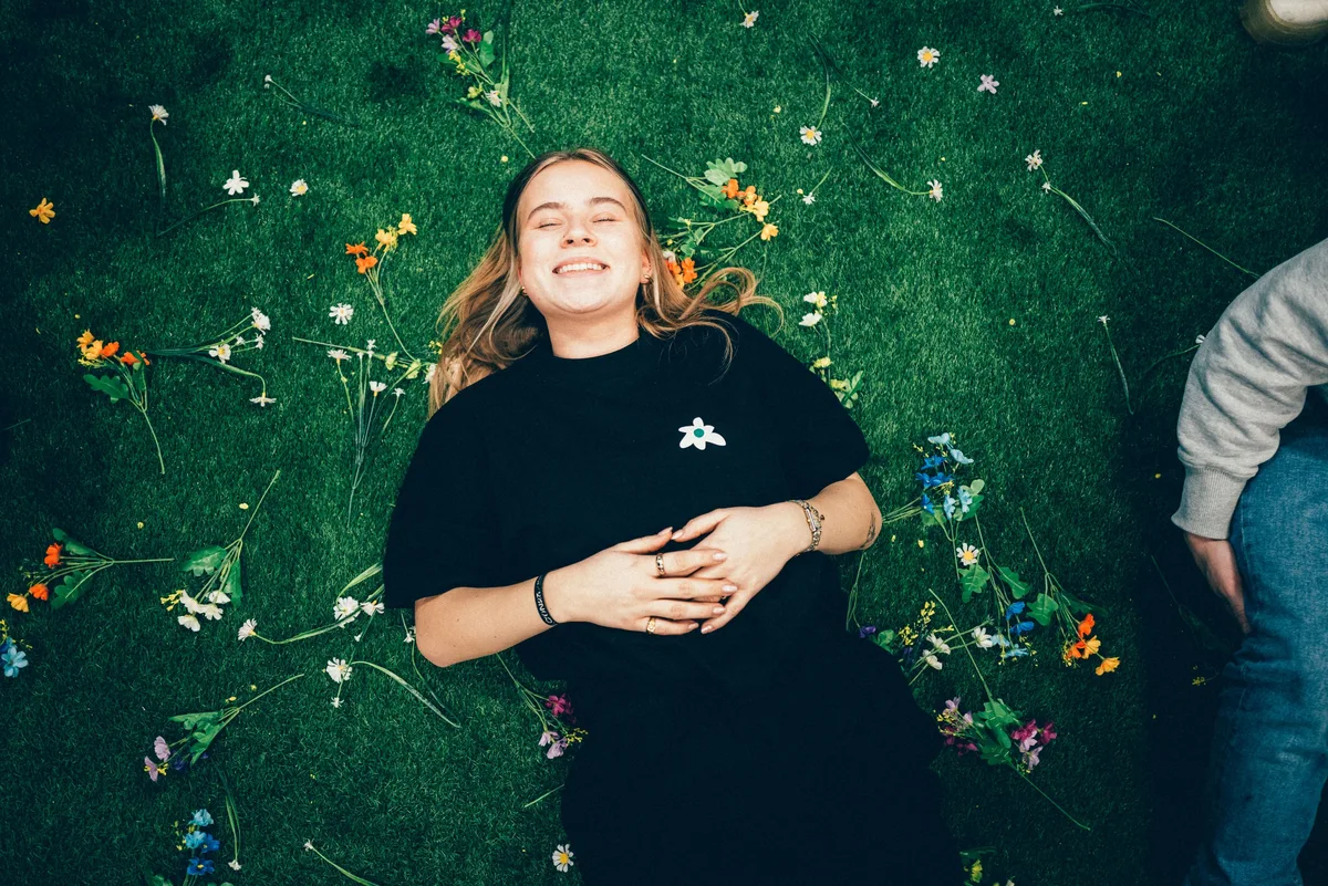 Draufsicht: Lachende Frau im schwarzen Shirt liegt mit geschlossenen Augen auf Kunstrasen zwischen verstreuten Blumen