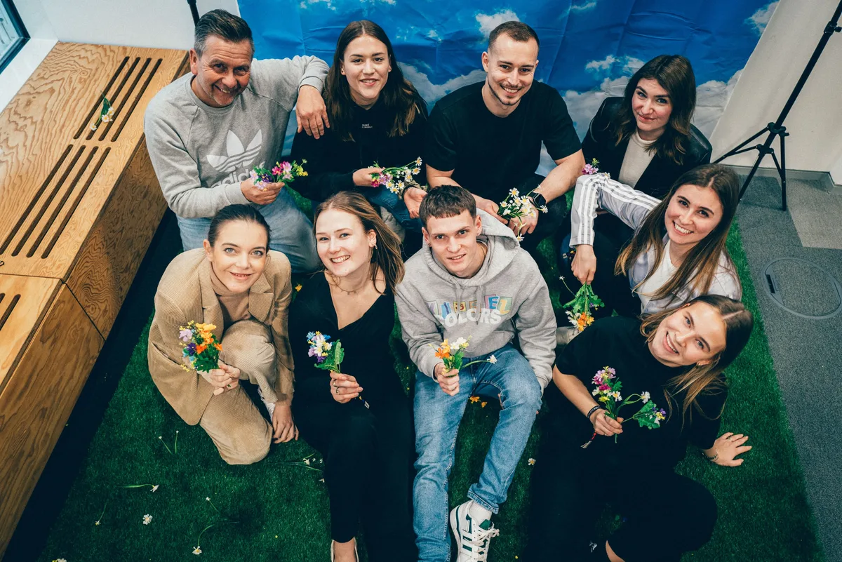 Draufsicht: Neun Menschen sitzen dicht auf Kunstrasen, jeder mit kleinem Blumenstrauß, alle blicken lächelnd nach oben