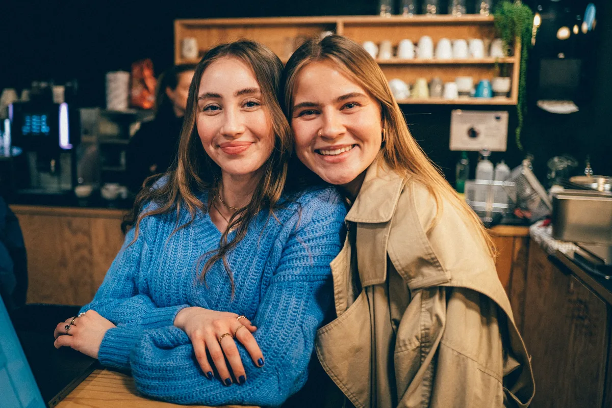 Dieselben beiden Frauen Kopf an Kopf, die eine im blauen Strickpulli, die andere im Trenchcoat, vor dem Küchenregal