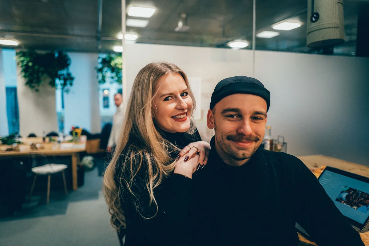 Blonde Frau lehnt lächelnd an einem Mann mit Mütze und Schnurrbart, rechts ein Laptop, links Holztische im Büro