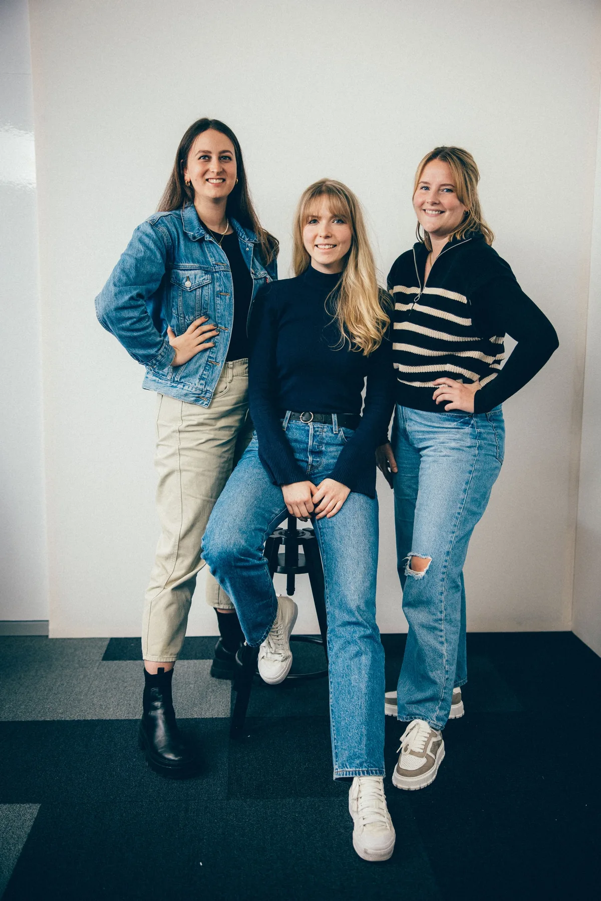 Drei Frauen vor weißer Wand im Hochformat, links in Jeansjacke, mittig auf dem Hocker im Rollkragen, rechts im Streifenpulli