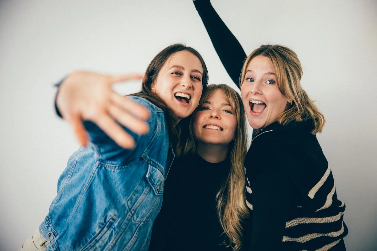 Dieselben drei Frauen lachen dicht beieinander, die linke streckt die Hand zur Kamera, rechts reißt eine den Mund auf