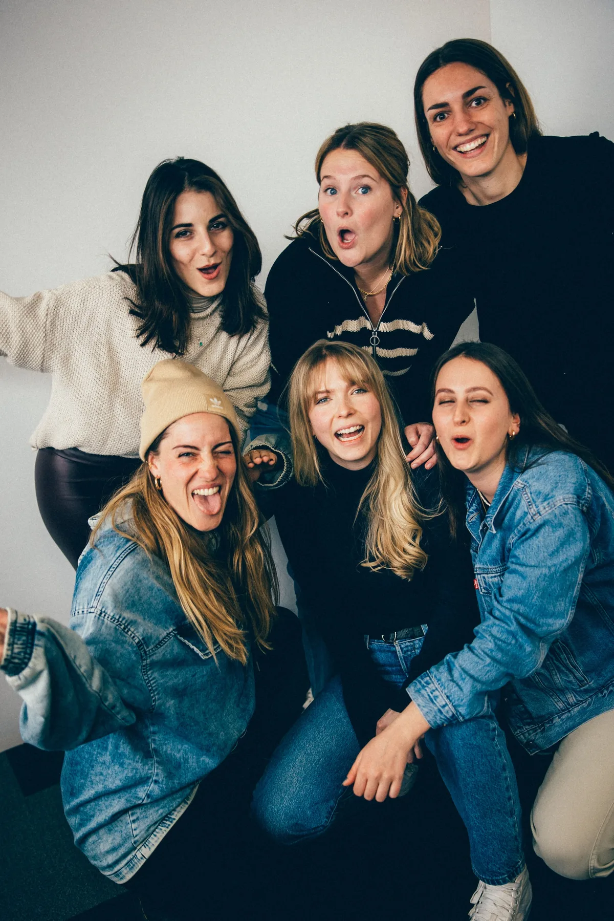 Sechs Frauen eng gedrängt vor weißer Wand im Hochformat, eine streckt die Zunge raus, eine zwinkert