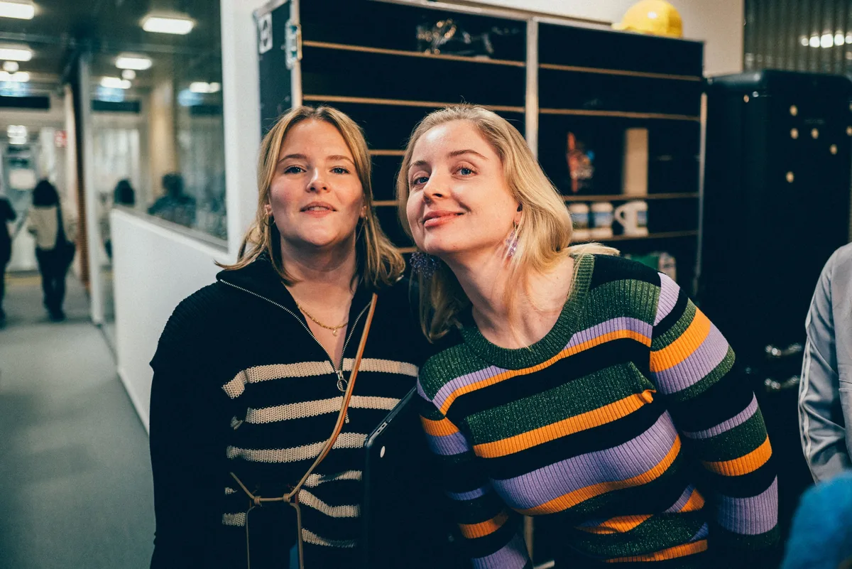 Zwei Frauen Kopf an Kopf, links im schwarz-weißen Zip-Pulli, rechts im bunt gestreiften Strickpulli, hinten ein Regal