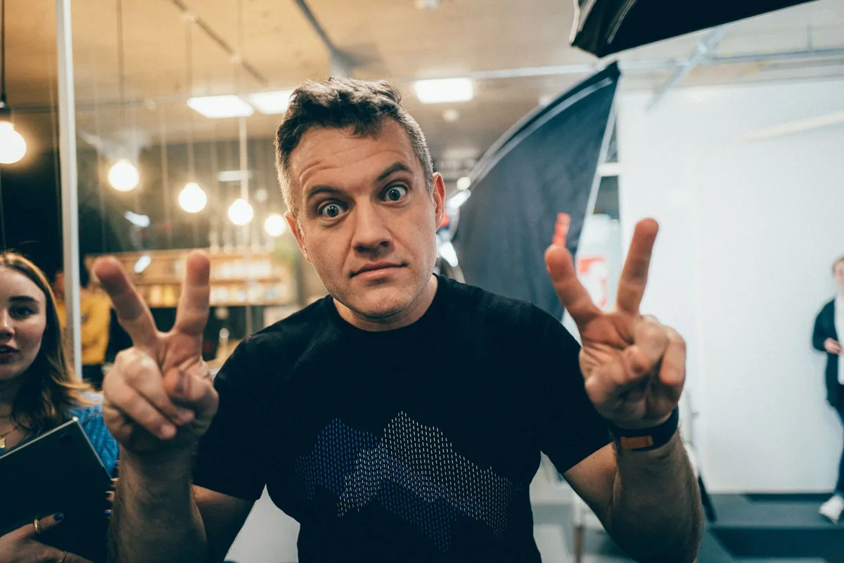 Mann im schwarzen T-Shirt reißt die Augen auf und hält beide Hände als Victory-Zeichen in die Kamera, links eine Softbox