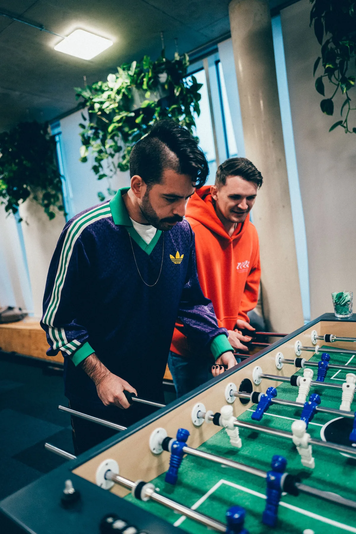 Zwei Männer am Kicker im Hochformat, einer im grün-violetten Trainingsshirt am Griff, der andere im orangen Hoodie schaut zu