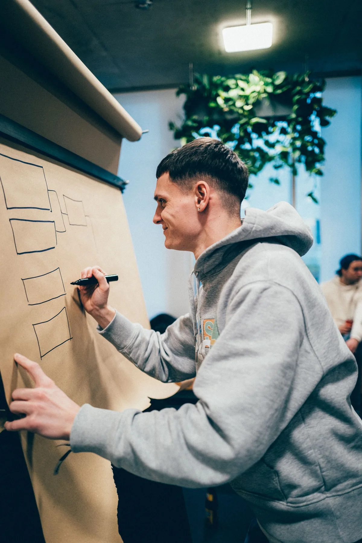 Mann im grauen Hoodie zeichnet mit Filzstift Kästen auf ein Flipchart-Papier, im Hintergrund Pflanzen und ein Fenster