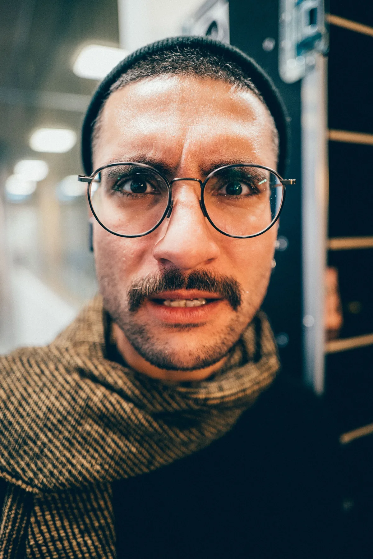 Sehr nahes Gesicht eines Mannes mit Mütze, Brille und Schnurrbart, er runzelt die Stirn, der Schal im Anschnitt