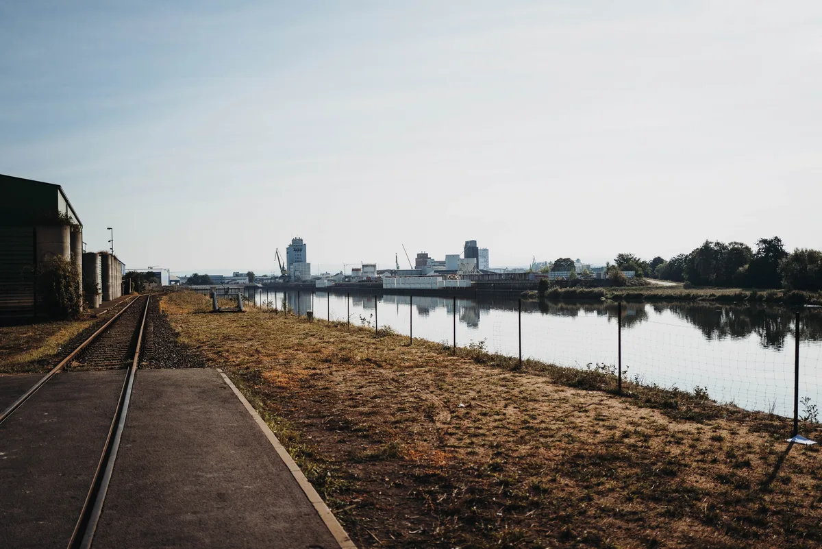 Gleis neben einer Asphaltfläche führt am Hafenbecken entlang, am anderen Ufer Silos und Kräne im Dunst