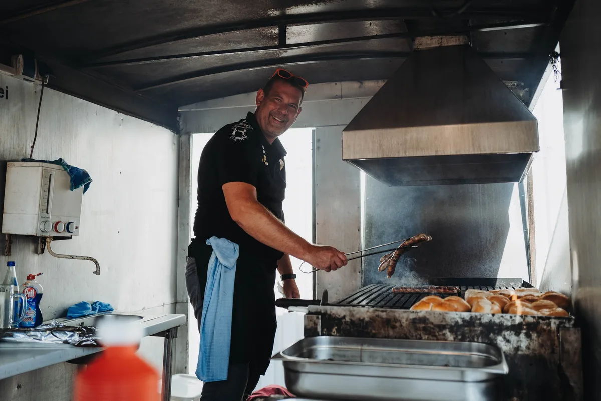 Lächelnder Mann in schwarzem Poloshirt und Schürze wendet in einem Grillwagen Bratwürste, daneben Brötchen auf dem Rost