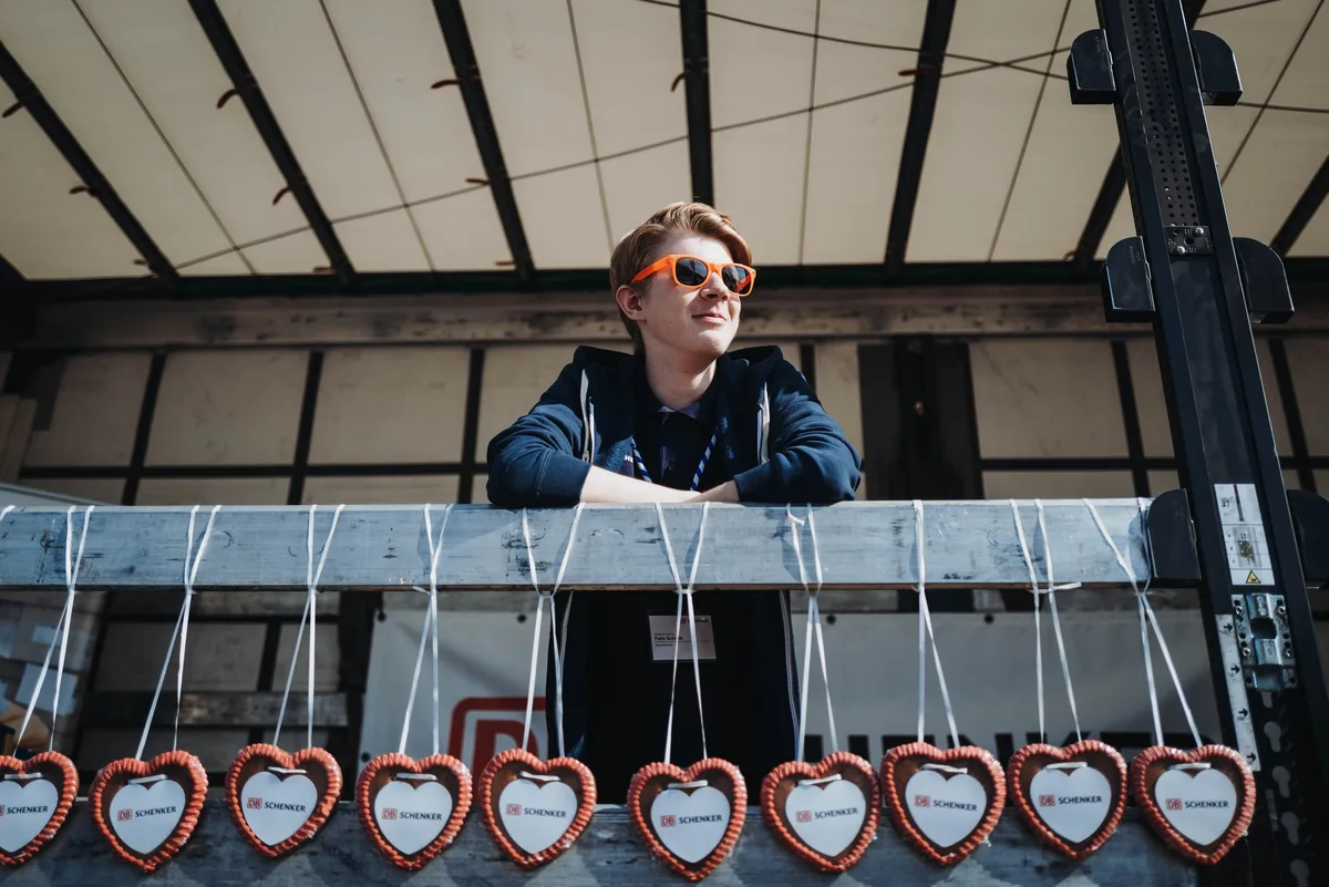 Junger Mann mit orangefarbener Sonnenbrille lehnt auf einer Ladefläche, vor ihm hängt eine Reihe Lebkuchenherzen