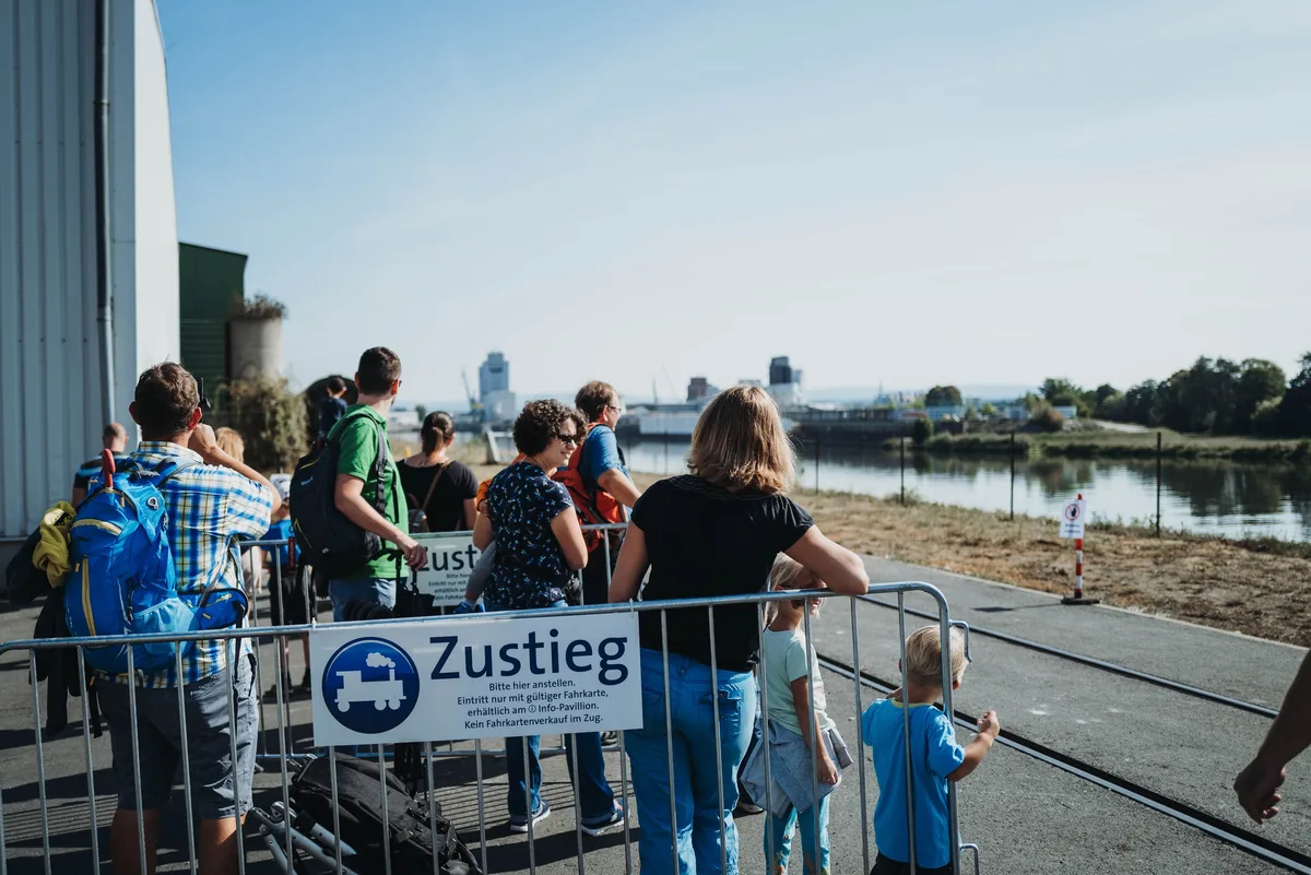Wartende Familien und Kinder stehen hinter einer Absperrung mit dem Schild „Zustieg“, dahinter das Hafenbecken