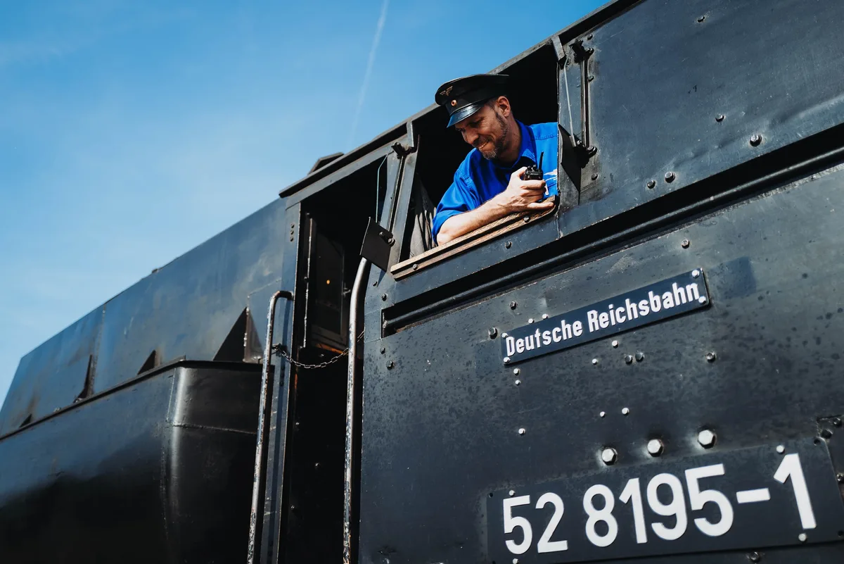 Lokführer mit Schirmmütze und blauem Hemd beugt sich aus dem Fenster einer schwarzen Dampflok, das Funkgerät in der Hand