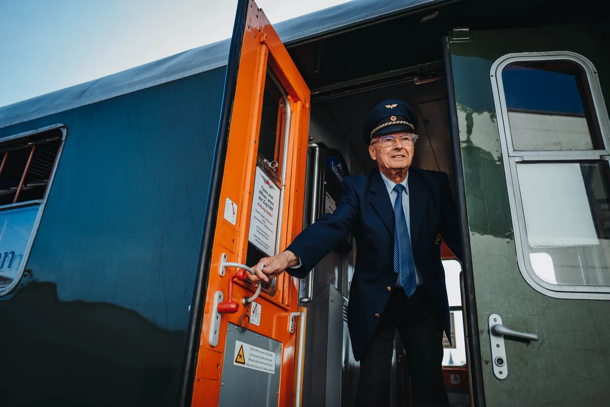 Älterer Schaffner in dunkler Uniform und Schirmmütze lehnt sich aus der orangefarbenen Wagentür und fasst den Haltegriff