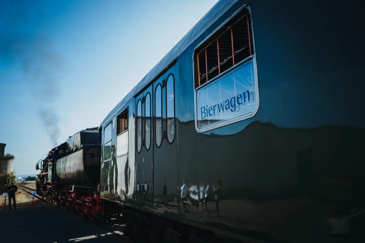 Der Bierwagen schräg von hinten im Gegenlicht, die Lok mit Rauchfahne im Hintergrund, links ein Besucher am Gleis
