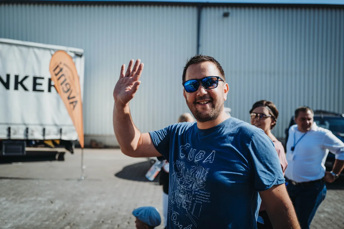 Mann mit blauer Spiegelsonnenbrille und Cuba-Shirt winkt lächelnd in die Kamera, hinter ihm Besucher vor einer Halle