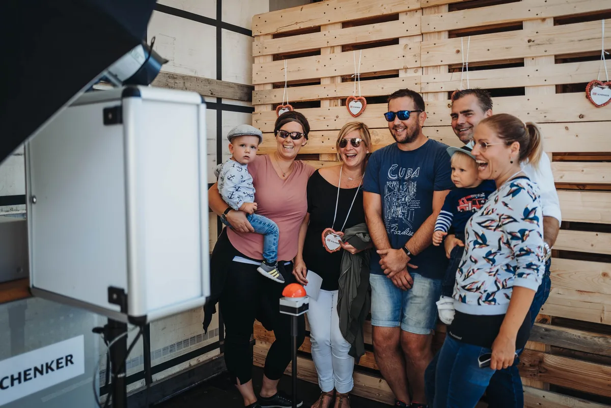 Zwei Paare mit zwei Kleinkindern posieren lachend vor einer Paletten-Wand neben einem Fotoauslöser mit roter Taste