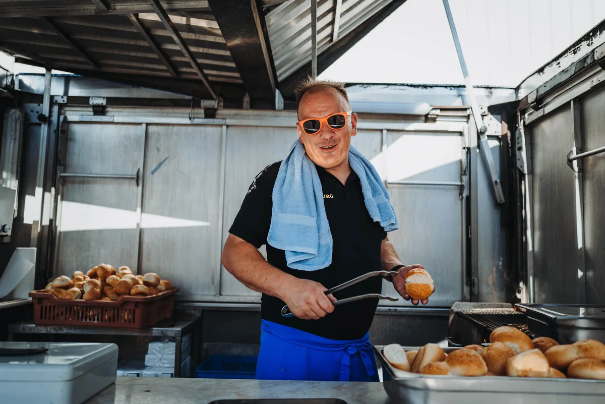 Koch mit orangefarbener Sonnenbrille und Handtuch um die Schultern hält mit der Zange ein Brötchen im Grillwagen