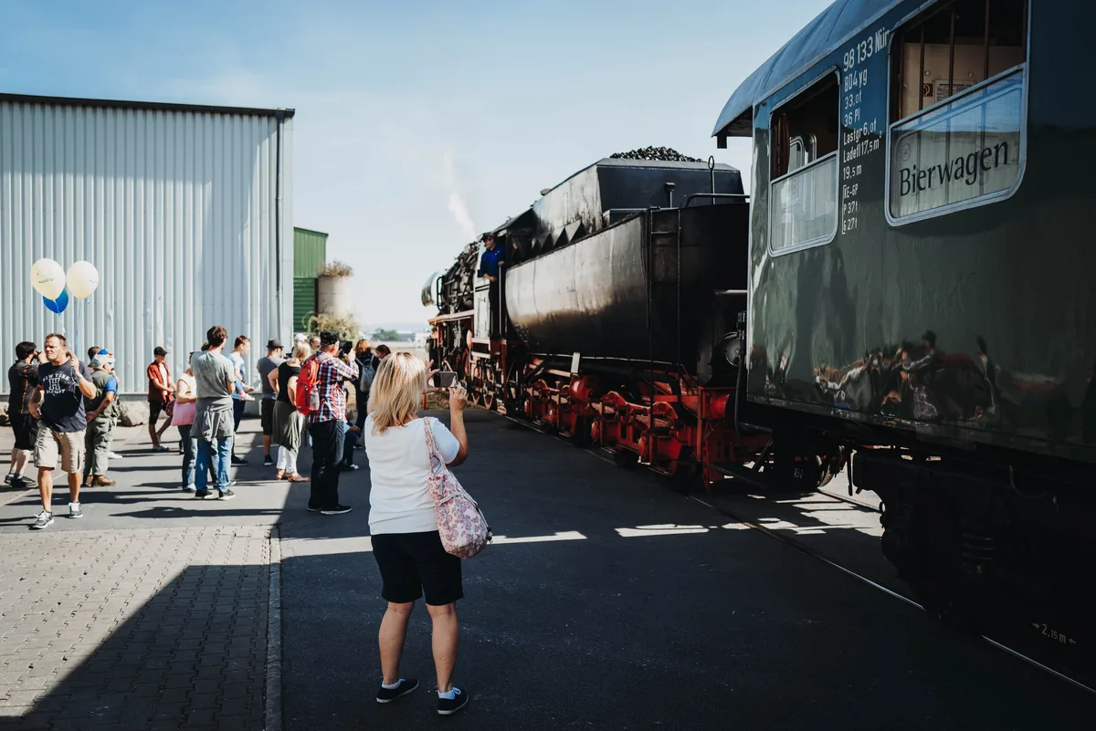 Besucher stehen am Gleis und fotografieren die einfahrende Dampflok, rechts der Bierwagen, links Luftballons vor der Halle