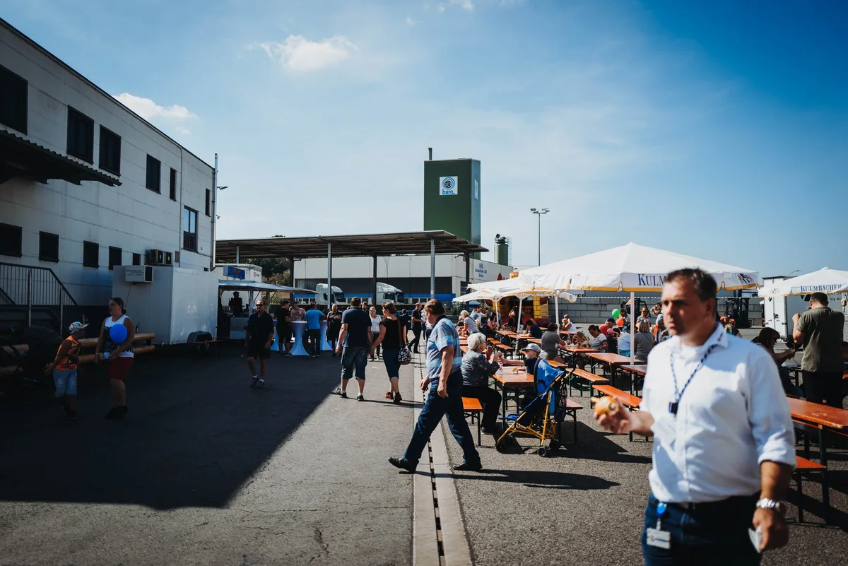 Hof mit Biertischgarnituren und Sonnenschirmen, Besucher laufen umher, vorn ein Mann im weißen Hemd mit Lanyard und Brötchen