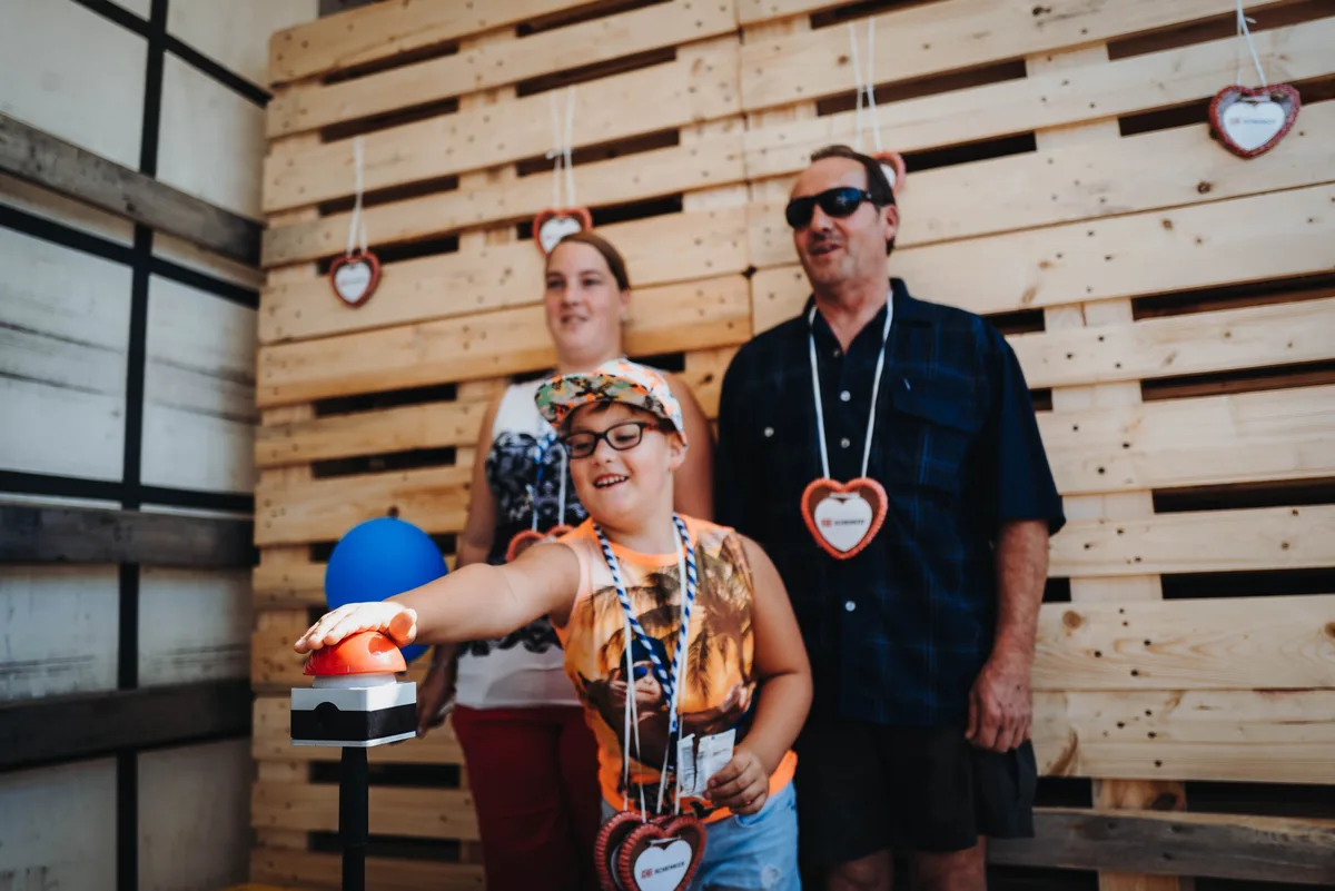 Kind mit Brille und Kappe drückt lachend auf den roten Fototaster, hinter ihm ein Paar vor einer Palettenwand