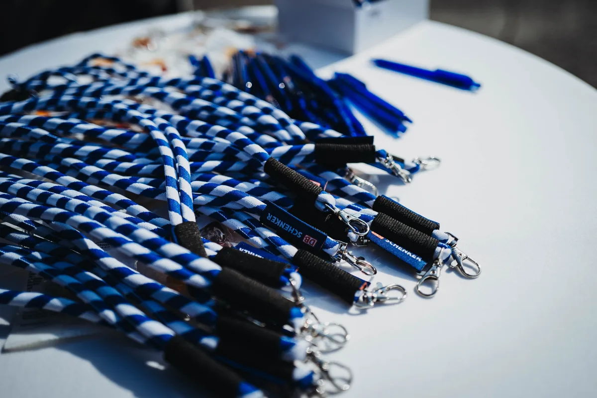 Blau-weiße Lanyards und blaue Kugelschreiber liegen auf einem weißen Stehtisch, an einigen hängt ein DB-Schenker-Anhänger