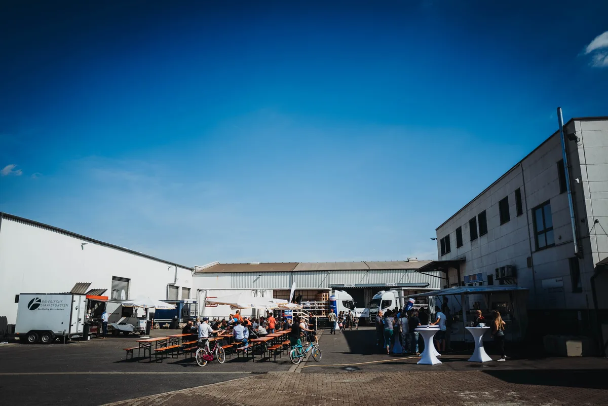 Weiter Blick über den Hof mit Biertischen, Imbisswagen und Stehtischen, rechts ein Gebäude, darüber tiefblauer Himmel
