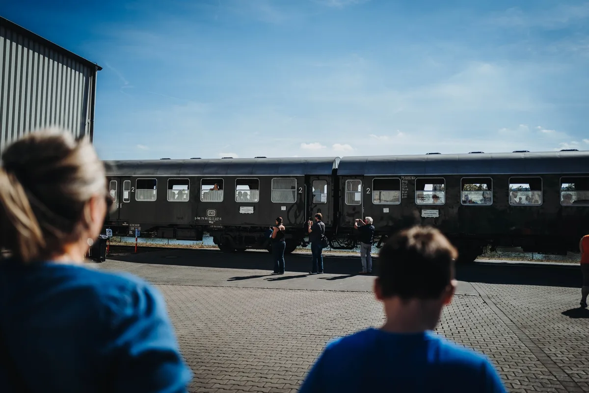Zug mit dunklen Personenwagen fährt am Hof vorbei, im Vordergrund unscharf ein Kind und eine Frau von hinten