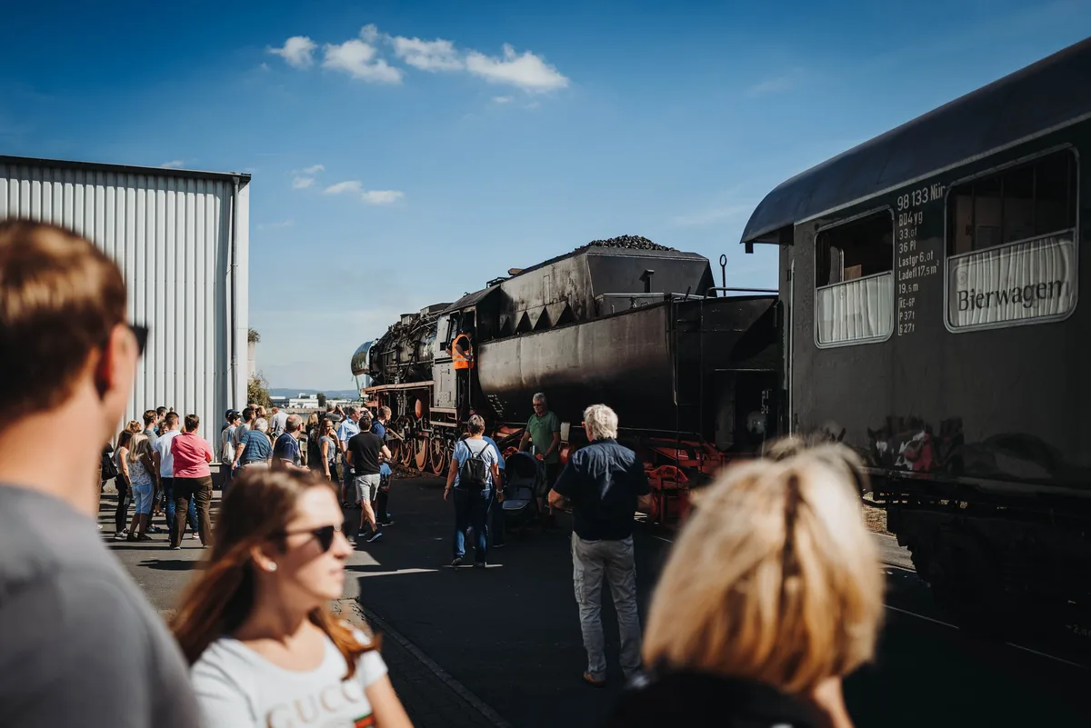 Besucher von hinten im Vordergrund, dahinter die Dampflok mit Kohletender und rechts der Bierwagen