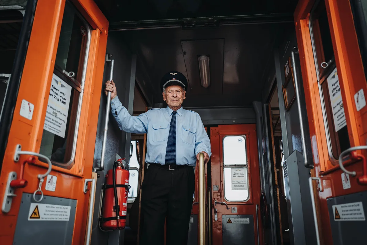 Älterer Schaffner steht in der Tür zwischen zwei orangefarbenen Wagentüren, eine Hand an der Stange, daneben ein Feuerlöscher